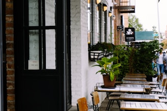 A cozy outdoor seating area with wooden tables and chairs positioned along a sidewalk. The exterior wall of the establishment is white brick, adorned with potted plants. A black-framed glass door with a 'Free WiFi' sign is visible. In the distance, a few people are walking, and there are hanging wall lamps providing ambient light.