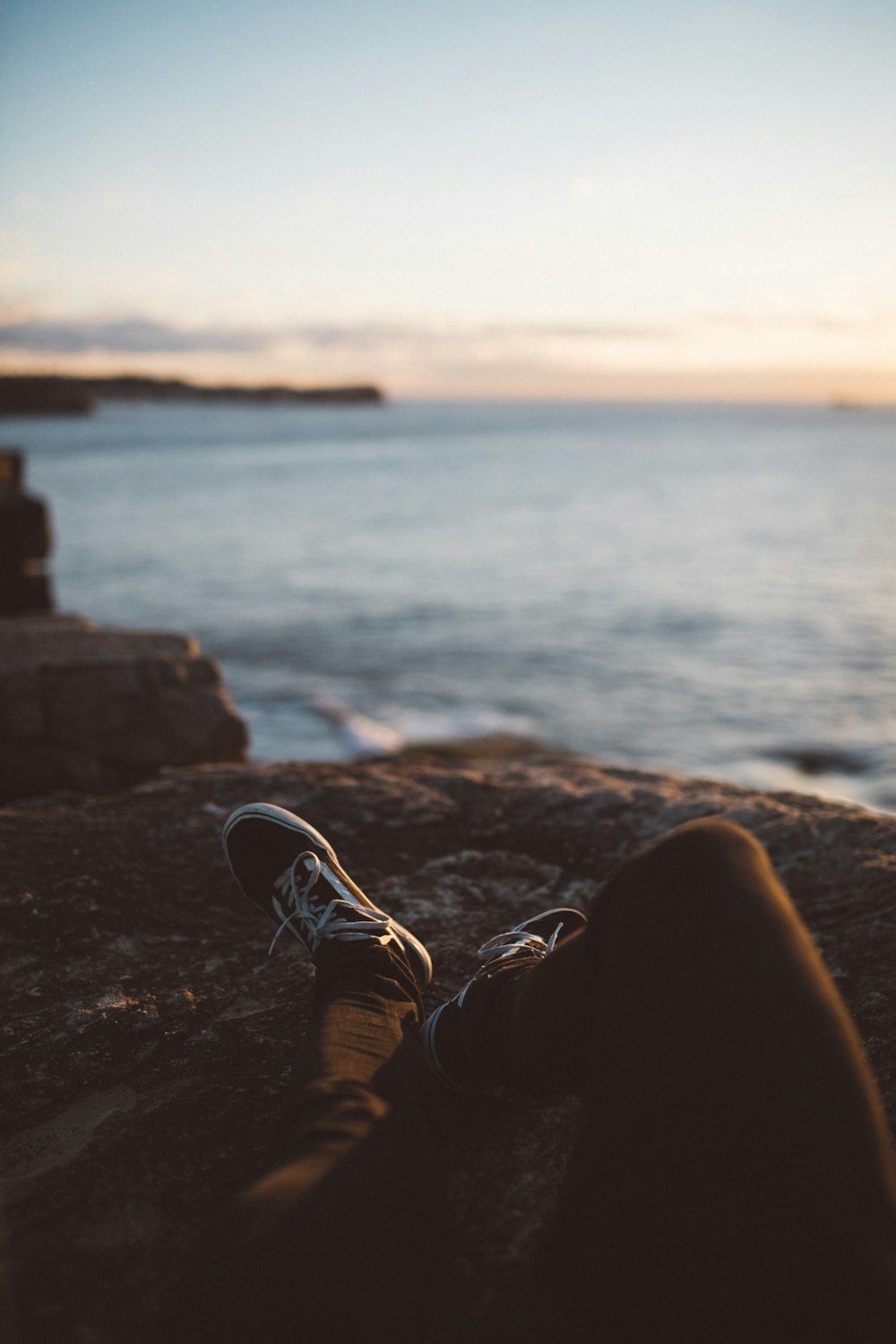 person wearing black pants and low-top sneakers lying down on brown rock formation near seashore