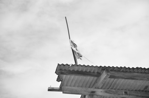 A corrugated metal roof with a wooden beam underneath. A tall pole extends upwards from the roof with a piece of fabric tied to it, fluttering in the wind. The sky is overcast with a light cloud cover.