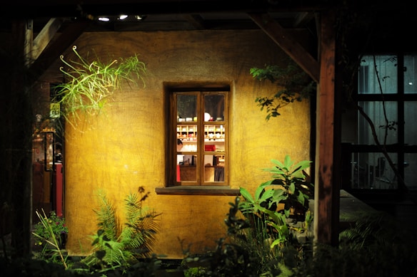A nighttime scene features a warmly lit rustic building facade with a central window. Through the window, shelves with various bottles are visible, creating a cozy ambience. The surrounding area includes lush green plants and foliage, adding a natural element to the setting.