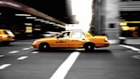 A yellow New York City taxi is speeding through an urban street, creating a sense of motion blur. The background features blurred cityscape, indicating movement and the hustle of city life.