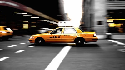 A yellow New York City taxi is speeding through an urban street, creating a sense of motion blur. The background features blurred cityscape, indicating movement and the hustle of city life.