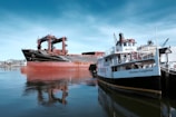 Two large ships are docked in calm waters near an industrial area. The ship on the left has a black hull with red and rust-colored sections, while the ship on the right, named Thomas Laighton, is white with black and yellow accents. The background shows cranes and industrial buildings.