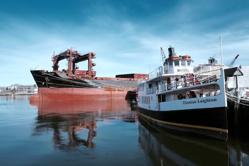 Two large ships are docked in calm waters near an industrial area. The ship on the left has a black hull with red and rust-colored sections, while the ship on the right, named Thomas Laighton, is white with black and yellow accents. The background shows cranes and industrial buildings.