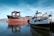 Two large ships are docked in calm waters near an industrial area. The ship on the left has a black hull with red and rust-colored sections, while the ship on the right, named Thomas Laighton, is white with black and yellow accents. The background shows cranes and industrial buildings.