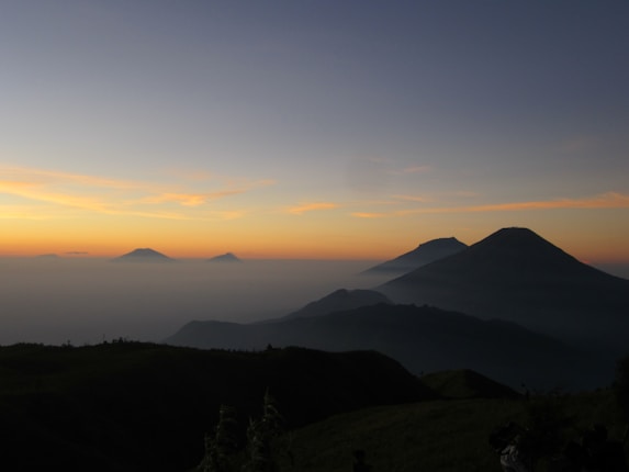 A serene mountain landscape at dawn with silhouetted peaks rising above a layer of mist. The sky displays a gradient of colors from blue to orange as the sun rises.