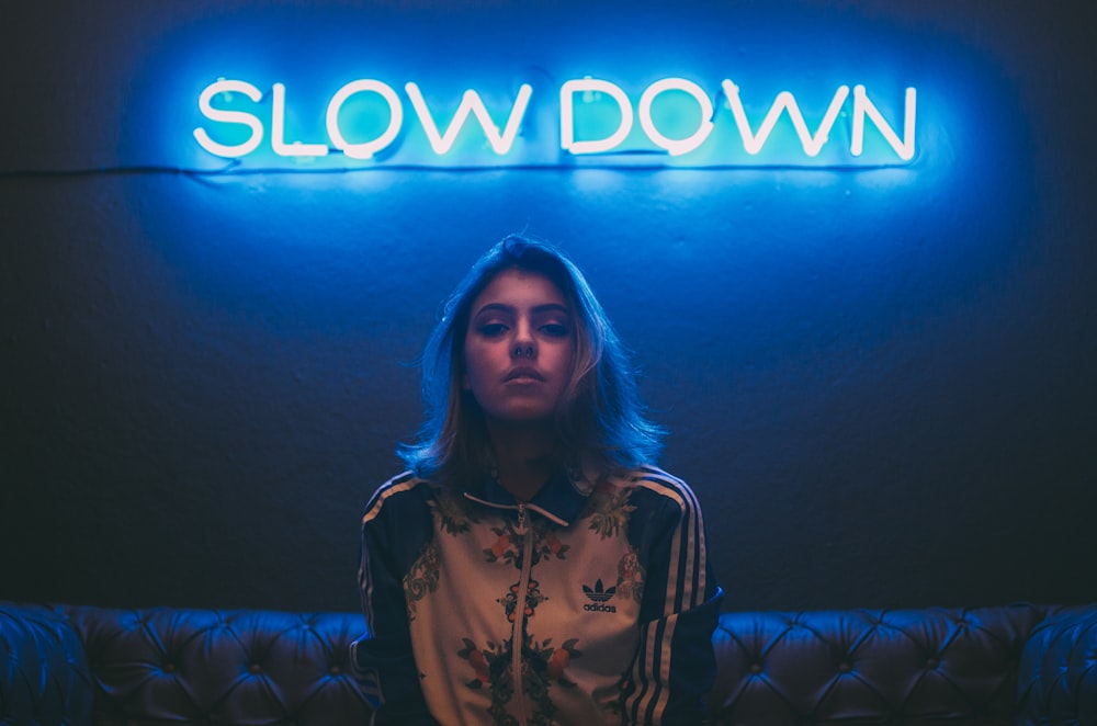 woman sitting on brown sofa under slow down neon signage