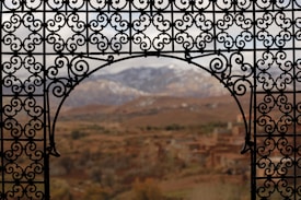 An ornate wrought iron balcony railing frames a scenic view of a mountain range in the distance, with a village nestled in the valley below. The ironwork features intricate scroll patterns that add a sense of elegance and craftsmanship.