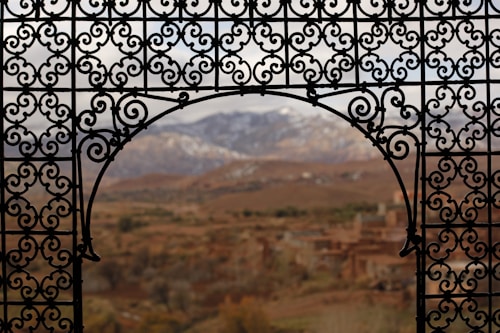 An ornate wrought iron balcony railing frames a scenic view of a mountain range in the distance, with a village nestled in the valley below. The ironwork features intricate scroll patterns that add a sense of elegance and craftsmanship.