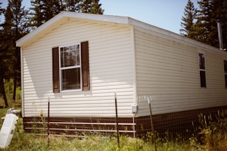 A mobile home with light-colored siding and dark window shutters sits surrounded by tall grass. A fence and some outdoor equipment are visible in front of the home, and there are tall evergreen trees in the background.