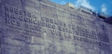 A stone or concrete wall with a large engraved inscription in Spanish. The text speaks about a task ordained by God and references Belgrano's proclamation. The wall features rough textures, and there are modern city buildings visible in the background, partially obscured by the wall.