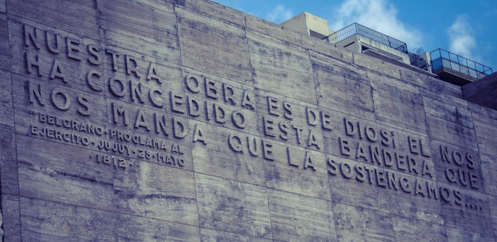 A stone or concrete wall with a large engraved inscription in Spanish. The text speaks about a task ordained by God and references Belgrano's proclamation. The wall features rough textures, and there are modern city buildings visible in the background, partially obscured by the wall.