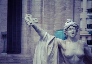 A stone statue depicts a bare-chested woman with a determined expression, holding a large cross aloft in her right hand. The background features a large stone building with steps leading up to a dark doorway, and part of a lantern possibly used for public lighting.