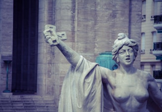 A stone statue depicts a bare-chested woman with a determined expression, holding a large cross aloft in her right hand. The background features a large stone building with steps leading up to a dark doorway, and part of a lantern possibly used for public lighting.