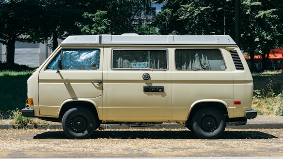 A vintage camper van parked on the side of a road surrounded by greenery. The van is beige with a gray roof and has curtains on the windows, suggesting it might be used for travel or camping. Behind the van, trees and buildings are partially visible.