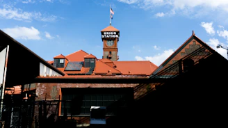 A red-brick building with a prominent tower showcasing a clock and a sign reading 'Union Station.' The roof has a series of red tiles, and an American flag flies atop the clock tower. The foreground features shadows and industrial elements, while the sky is clear with a few scattered clouds.