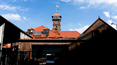 A red-brick building with a prominent tower showcasing a clock and a sign reading 'Union Station.' The roof has a series of red tiles, and an American flag flies atop the clock tower. The foreground features shadows and industrial elements, while the sky is clear with a few scattered clouds.