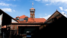 A red-brick building with a prominent tower showcasing a clock and a sign reading 'Union Station.' The roof has a series of red tiles, and an American flag flies atop the clock tower. The foreground features shadows and industrial elements, while the sky is clear with a few scattered clouds.