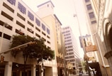 An urban street scene features tall commercial buildings with various architectural styles, including modern and classical elements. Flags of Texas and the United States are displayed. Trees line the sidewalk alongside street signs directing towards popular locations like the Alamo and a theater. The street is relatively quiet, with minimal traffic and activity.