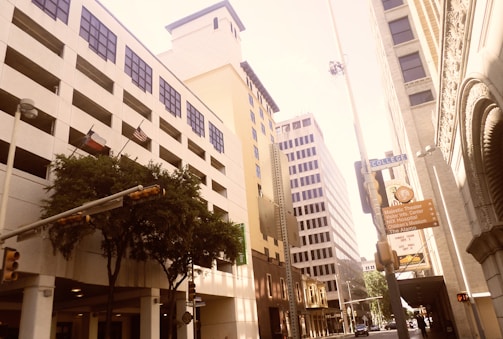An urban street scene features tall commercial buildings with various architectural styles, including modern and classical elements. Flags of Texas and the United States are displayed. Trees line the sidewalk alongside street signs directing towards popular locations like the Alamo and a theater. The street is relatively quiet, with minimal traffic and activity.