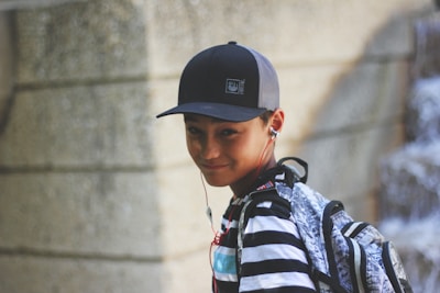 A young person wearing a black cap and striped shirt is listening to music with earbuds. They are carrying a backpack, and there is a slight smile on their face. The background consists of a textured stone wall and soft focus on what may be a waterfall or flowing water.