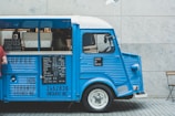 A vintage blue coffee truck is parked with its side window open, displaying a menu with various coffee options. The truck is stationed against a light stone wall, and part of a wooden chair is visible to the right. A person wearing a red shirt stands to the left, partially obscured. The truck features signs with the name 'Jacked Up Coffee' and has several details like its license number and location, Toronto, printed on it.