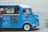 A vintage blue coffee truck is parked with its side window open, displaying a menu with various coffee options. The truck is stationed against a light stone wall, and part of a wooden chair is visible to the right. A person wearing a red shirt stands to the left, partially obscured. The truck features signs with the name 'Jacked Up Coffee' and has several details like its license number and location, Toronto, printed on it.