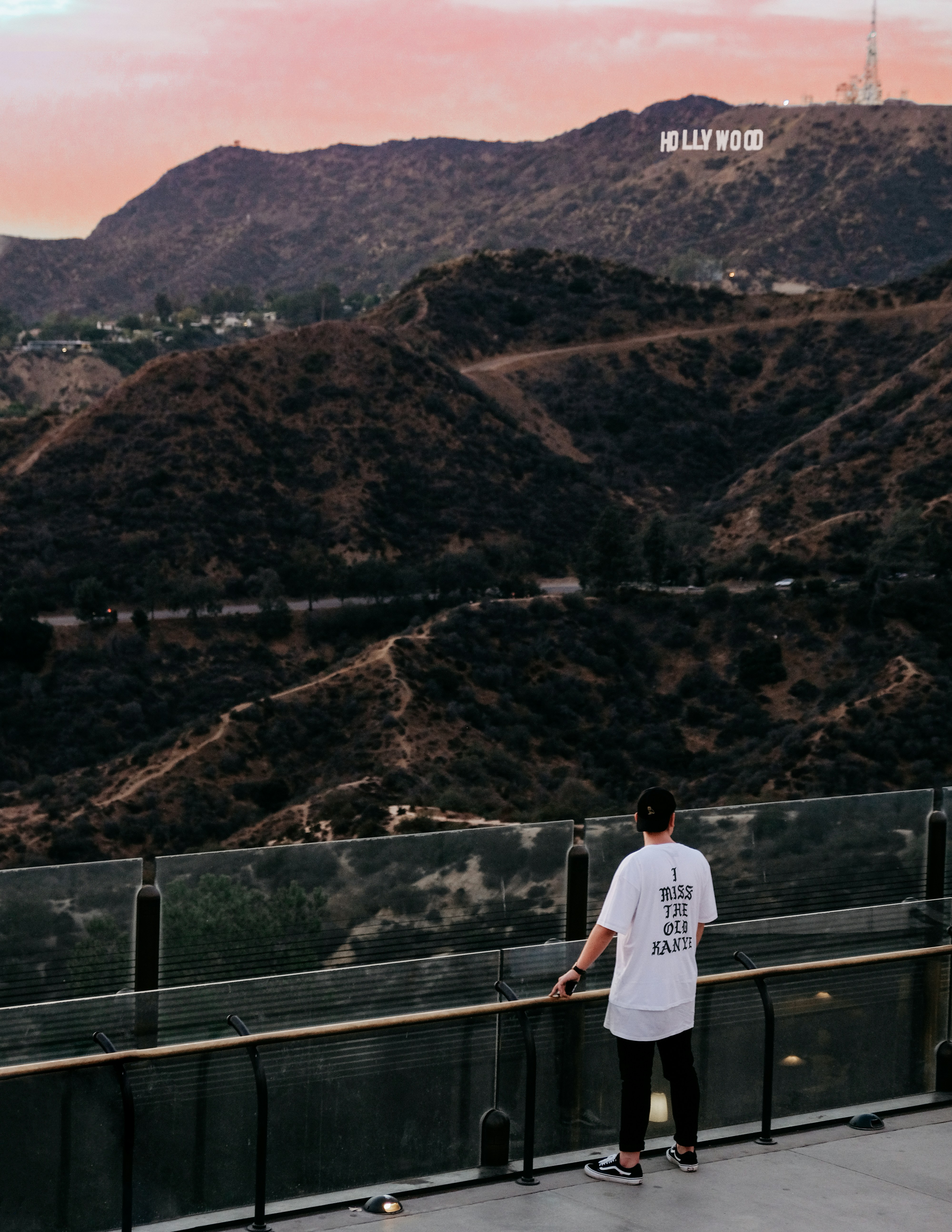 A solitary figure leans on a glass railing at a lookout, gazing over rugged hills. The distant Hollywood Sign sits atop the hills against a pink sunset.