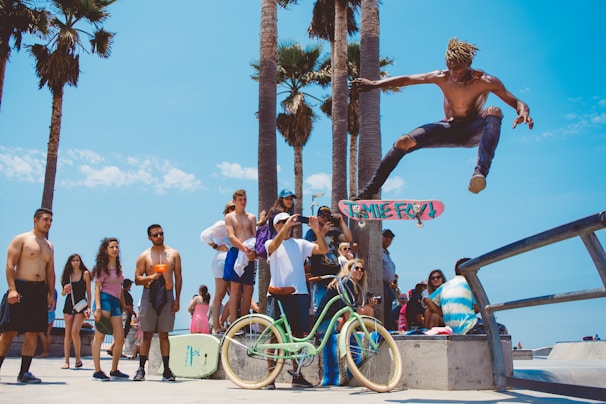 A skateboarder performs a high jump trick in mid-air at a skate park near the beach. Several onlookers, including men and women in casual summer clothing, watch the action from nearby. Palm trees are in the background, and a bright blue sky with a few clouds adds to the summery feel. Bicycles and surfboards are also visible.
