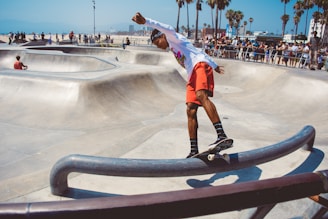 A skateboarder wearing a white long-sleeve shirt and red shorts balances on a skateboard while grinding along a metal rail in an outdoor skate park. Surrounding him are smooth concrete ramps and bowls, with palm trees visible in the background. A crowd of people watches the action, and a person sits casually on one of the ramps.