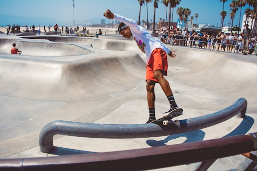 A skateboarder wearing a white long-sleeve shirt and red shorts balances on a skateboard while grinding along a metal rail in an outdoor skate park. Surrounding him are smooth concrete ramps and bowls, with palm trees visible in the background. A crowd of people watches the action, and a person sits casually on one of the ramps.