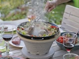 A backyard dining scene with a variety of vegetables grilling on a stone grill, while wine glasses and fresh ingredients are placed on a wooden table. A hand holds tongs, tending to the food, suggesting an outdoor gathering.