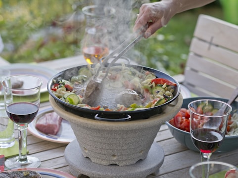 A backyard dining scene with a variety of vegetables grilling on a stone grill, while wine glasses and fresh ingredients are placed on a wooden table. A hand holds tongs, tending to the food, suggesting an outdoor gathering.