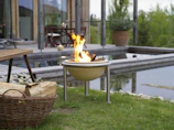 A serene outdoor setting features a small, round fire pit with flames burning brightly on a grassy lawn. A wicker basket filled with firewood rests nearby. In the background, a wooden deck with patio furniture overlooks a modern rectangular water feature bordered by stones. Lush greenery surrounds the area, adding to the tranquil atmosphere.