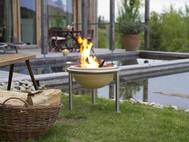 A serene outdoor setting features a small, round fire pit with flames burning brightly on a grassy lawn. A wicker basket filled with firewood rests nearby. In the background, a wooden deck with patio furniture overlooks a modern rectangular water feature bordered by stones. Lush greenery surrounds the area, adding to the tranquil atmosphere.
