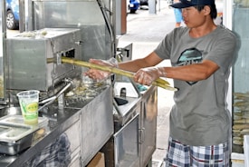 A person operates a sugar cane juicing machine, feeding a stalk into the metal apparatus. The individual wears gloves and a cap, focusing on the task. The setup appears to be part of a street-side or market stall, with a clear cup containing a green beverage nearby.
