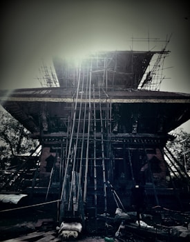 The image depicts a large, ancient temple structure under renovation, with scaffolding and construction materials surrounding it. The structure has a tiered roof, supported by wooden poles, and intricate carvings on the walls are visible. There is a bright light source at the top center, possibly the sun, creating a hazy aura around the temple. A solitary figure is seen at the lower right corner, adding a sense of scale.