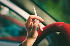 A hand is holding a lit cigarette, with fingers resting on a red steering wheel. The image is taken from inside a vehicle, with blurred green and red hues visible through the window.