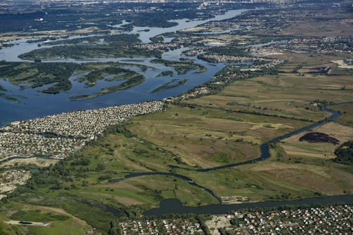 An aerial view of a landscape featuring a wide river winding through lush green fields and patches of forest. Numerous settlements with clustered houses are visible along the riverbanks. The landscape extends into the distance with diverse vegetation surrounding the water bodies.