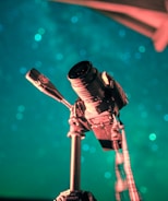 A camera mounted on a tripod is set against a backdrop of teal and blue bokeh lights. The angle suggests a focus on capturing the night sky. The lighting on the camera highlights its structure and details, creating an artistic and professional atmosphere.