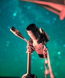 A camera mounted on a tripod is set against a backdrop of teal and blue bokeh lights. The angle suggests a focus on capturing the night sky. The lighting on the camera highlights its structure and details, creating an artistic and professional atmosphere.