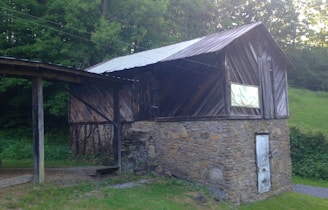A rustic wooden barn with a stone foundation stands surrounded by lush green foliage. The roof is metal, and a wooden door is visible at the stone level. A sign with faded writing is affixed to the barn's upper section.