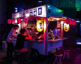 A vibrant food truck adorned with colorful lanterns serves customers at night. Two people sit at the counter, illuminated by the soft, warm glow of hanging lanterns. The setting has a cozy and inviting ambiance as the neon lights create dynamic reflections on the truck.
