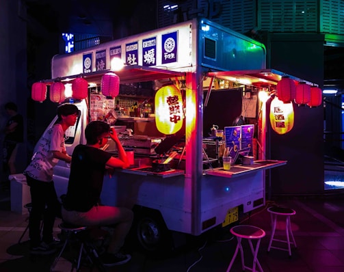 A vibrant food truck adorned with colorful lanterns serves customers at night. Two people sit at the counter, illuminated by the soft, warm glow of hanging lanterns. The setting has a cozy and inviting ambiance as the neon lights create dynamic reflections on the truck.