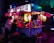 A vibrant food truck adorned with colorful lanterns serves customers at night. Two people sit at the counter, illuminated by the soft, warm glow of hanging lanterns. The setting has a cozy and inviting ambiance as the neon lights create dynamic reflections on the truck.