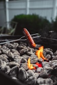 A sausage being cooked over a bed of hot charcoal. The charcoals are glowing with a vibrant flame, indicating they are exceptionally hot. The surrounding environment appears to be an outdoor setting with a blurred background.