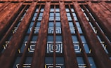 An architectural facade features tall, vertically oriented columns made of brown stone that frame rows of large, reflective glass windows. The glass windows display geometric patterns created by illuminated yellow and white lines.
