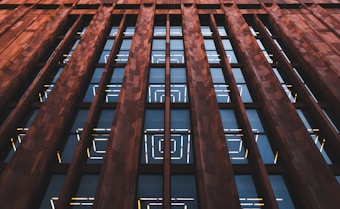 An architectural facade features tall, vertically oriented columns made of brown stone that frame rows of large, reflective glass windows. The glass windows display geometric patterns created by illuminated yellow and white lines.