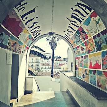 A beautifully painted archway features colorful comic-style artwork, with scenes depicted in vibrant panels on the walls. Through the archway, a view of a cityscape with quaint buildings and a tall palm tree is visible. The setting appears charming and artistic.