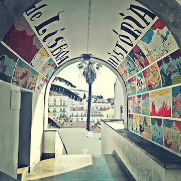 A beautifully painted archway features colorful comic-style artwork, with scenes depicted in vibrant panels on the walls. Through the archway, a view of a cityscape with quaint buildings and a tall palm tree is visible. The setting appears charming and artistic.